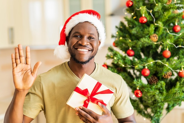 Hombre afroamericano que se siente feliz porque recibe un regalo de navidad y saluda a la cámara y dice hola