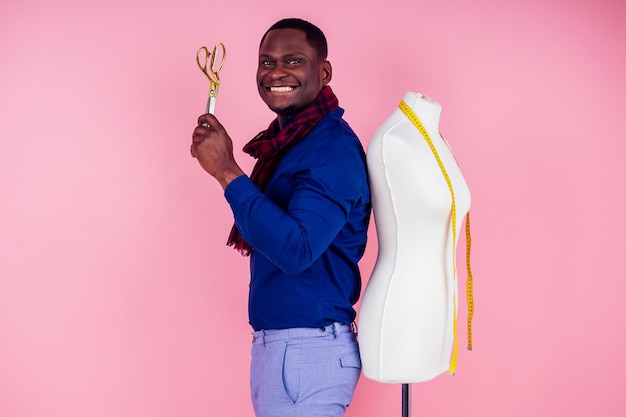 Hombre afroamericano sastre costurera taller elegante modelo masculino diseñador de ropa cinta métrica en el cuello posando junto al maniquí sosteniendo tijeras en la mano sobre fondo rosa en el estudio