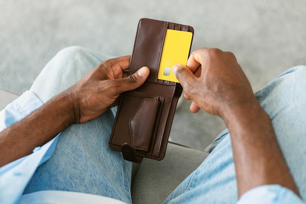 Hombre afroamericano seleccionando tarjeta de crédito de su billetera sentado en el sofá en casa cerrado