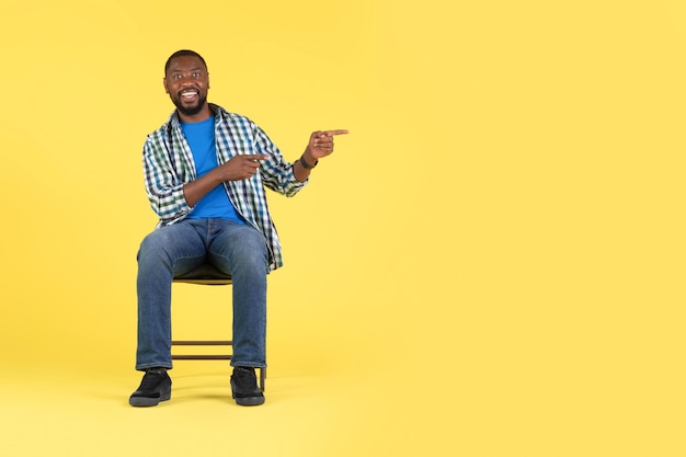 Hombre afroamericano señalando con el dedo a un lado sentado sobre fondo amarillo