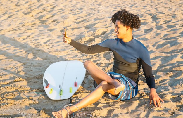 Hombre afroamericano sentado en la arena con tabla de surf toma una foto con un teléfono inteligente