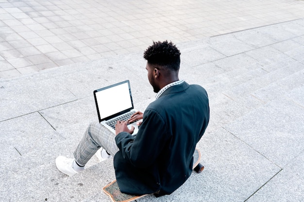 Hombre afroamericano sentado en las escaleras de la ciudad de espaldas usando su laptop para trabajar