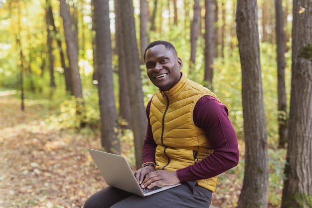 Hombre afroamericano sentado en el parque de la ciudad en un banco con una computadora portátil estudiando en línea al aire libre