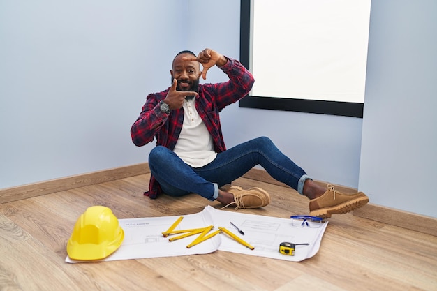 Hombre afroamericano sentado en el suelo en un nuevo hogar mirando planos sonriendo haciendo marco con manos y dedos con cara feliz. concepto de creatividad y fotografía.