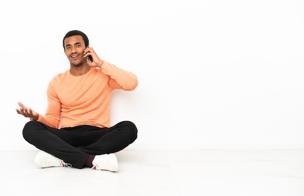 Hombre afroamericano sentado en el suelo sobre fondo copyspace aislado manteniendo una conversación con el teléfono móvil con alguien