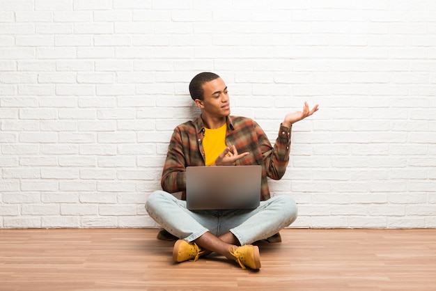 Hombre afroamericano sentado en el suelo con su computadora portátil extendiendo las manos hacia un lado para invitar a venir