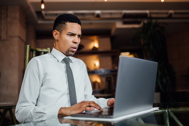 Hombre afroamericano seriamente sorprendido por lo que ve en su computadora portátil, con una camisa blanca, interior