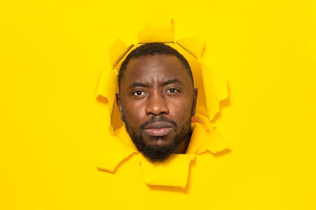 Un hombre afroamericano serio posando en un fondo de papel amarillo roto mirando la cámara a través del agujero
