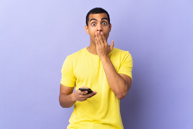 Hombre afroamericano sobre pared púrpura aislado mediante teléfono móvil con expresión facial sorpresa