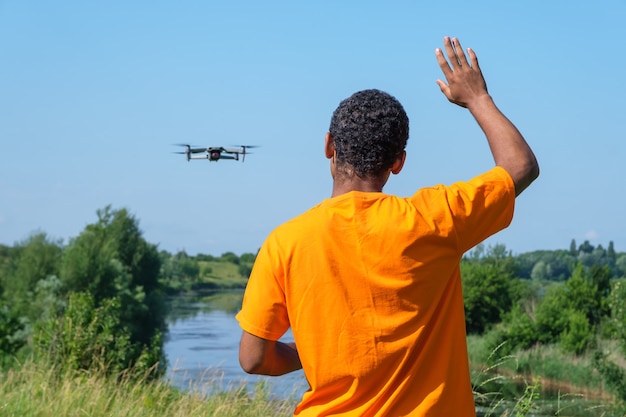 Hombre afroamericano sonriente joven que opera drone con controlador de pie con la espalda y saludando con la mano en el prado cerca del río.