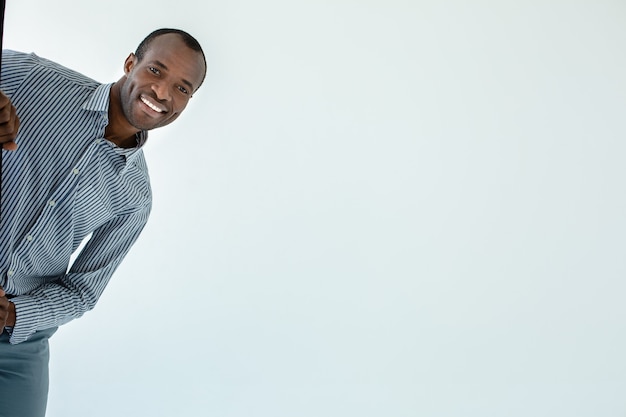 Hombre afroamericano sonriente positivo que se siente feliz mientras se esconde contra la pared blanca