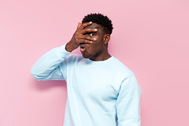 Un hombre afroamericano sorprendido con un suéter azul se cubre la cara con las manos escondiéndose y mirando