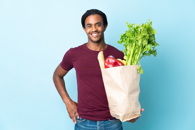 Hombre afroamericano sosteniendo una bolsa de compras aislada en azul posando con los brazos en la cadera y sonriendo