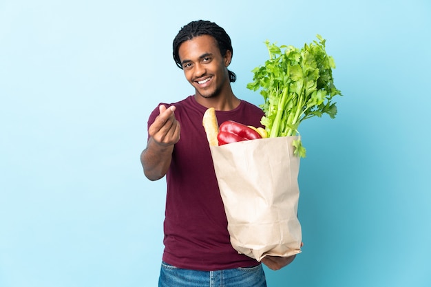 Hombre afroamericano sosteniendo una bolsa de compras aislada en la pared azul haciendo gesto de dinero