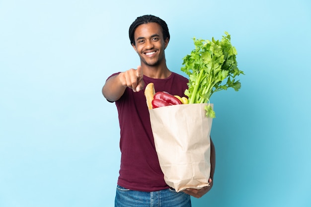 Hombre afroamericano sosteniendo una bolsa de compras aislado sobre fondo azul apuntando hacia el frente con expresión feliz