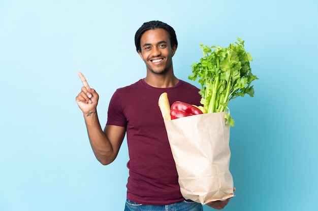 Hombre afroamericano sosteniendo una bolsa de compras aislado sobre fondo azul mostrando y levantando un dedo en señal de lo mejor