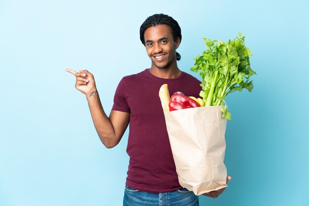 Hombre afroamericano sosteniendo una bolsa de compras aislado sobre fondo azul que señala el dedo hacia el lado