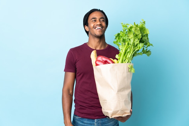 Hombre afroamericano sosteniendo una bolsa de compras aislado sobre fondo azul riendo