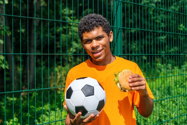 Hombre afroamericano sosteniendo una pelota de fútbol mientras está de pie contra la valla