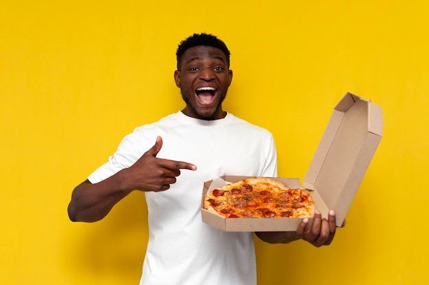 hombre afroamericano sostiene una caja de pizza y la señala con el dedo en el fondo amarillo