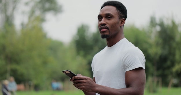 Hombre afroamericano con teléfono inteligente dentro del parque de la ciudad después de hacer ejercicio al aire libre. Hombre de fitness atleta mensajes de texto mirando la pantalla del teléfono después de hacer ejercicio.