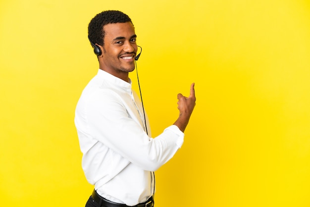 Hombre afroamericano Telemarketer trabajando con un auricular sobre fondo amarillo aislado apuntando hacia atrás