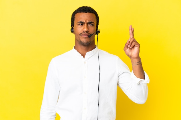 Hombre afroamericano Telemarketer trabajando con un auricular sobre fondo amarillo aislado con los dedos cruzando y deseando lo mejor