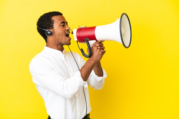 Hombre afroamericano Telemarketer trabajando con un auricular sobre fondo amarillo aislado gritando a través de un megáfono