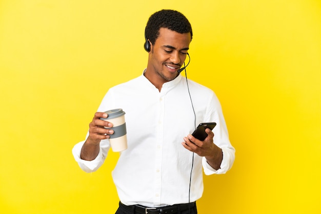 Hombre afroamericano Telemarketer trabajando con un auricular sobre fondo amarillo aislado sosteniendo café para llevar y un móvil