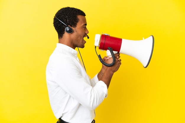 Hombre afroamericano Telemarketer trabajando con un auricular sobre pared amarilla aislada gritando a través de un megáfono