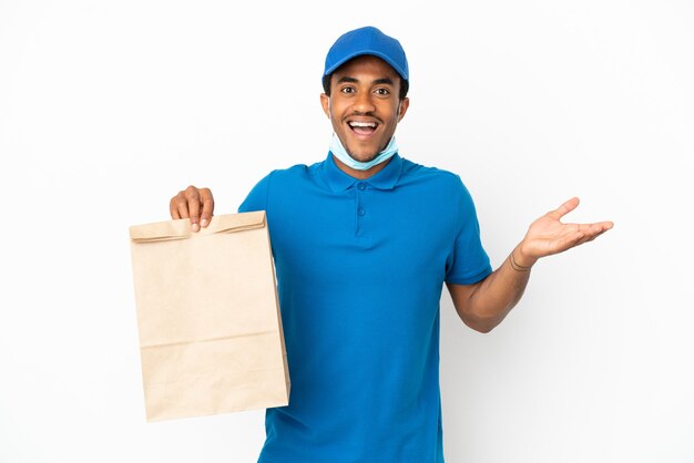 Hombre afroamericano tomando una bolsa de comida para llevar aislado sobre fondo blanco con expresión facial sorprendida