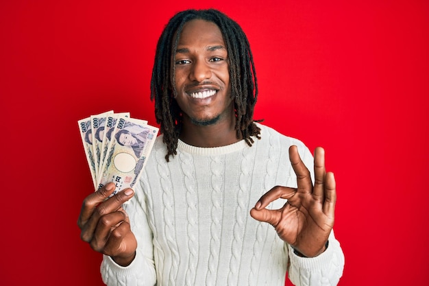 Hombre afroamericano con trenzas sosteniendo billetes de 5000 yenes japoneses haciendo un buen signo con los dedos sonriendo amigablemente gesticulando excelente símbolo