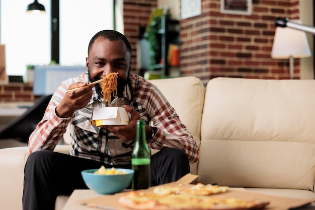 Hombre afroamericano usando palillos para comer comida de fideos para llevar en casa, viendo películas en el canal de televisión. Comer comida para llevar de la caja y disfrutar de la película en la televisión.