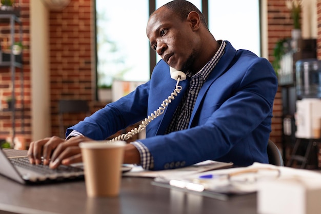 Hombre afroamericano usando teléfono fijo para conversar sobre estrategia de negocios. Empresario hablando por teléfono con el gerente para planificar un proyecto para un servicio exitoso en la oficina.