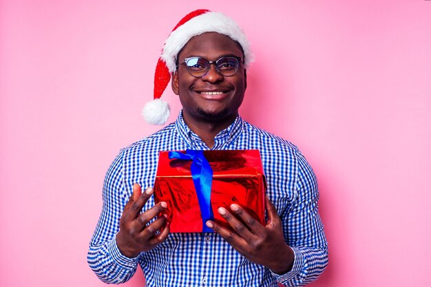 Hombre afroamericano vistiendo elegante camisa a cuadros gran sonrisa en santa sombrero con caja de regalo sobre fondo rosa studio.Papá Noel de piel oscura felicitando feliz navidad