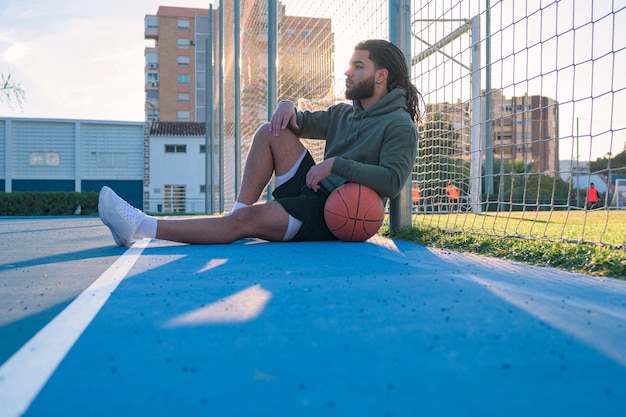 Hombre afrolatino con una pelota sentada en una cancha de baloncesto al atardecer