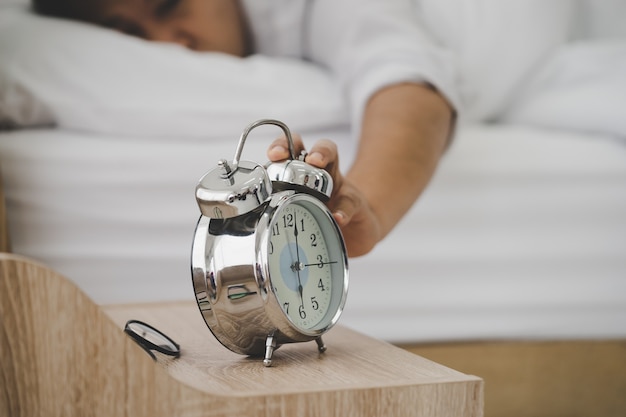 El hombre agotado que duerme en la cama está cansado al despertarse con el timbre del despertador en la mañana que sufre de insomnio.