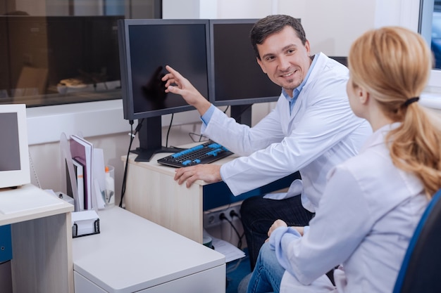 Hombre agradable sonriente positivo apuntando a la pantalla del ordenador y mirándola mientras habla con ella