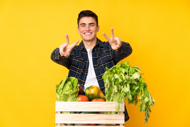 Hombre agricultor adolescente con verduras recién cortadas en una caja sonriendo y mostrando el signo de la victoria