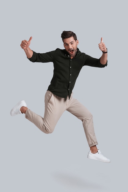 Hombre en el aire. Longitud total de joven juguetón gesticulando y sonriendo mientras salta contra el fondo gris