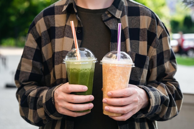 Un hombre al aire libre sostiene vasos de plástico con batidos verdes y naranjas, una deliciosa bebida refrescante de verano