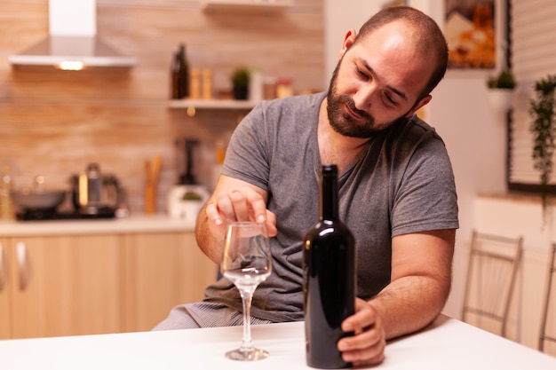 Foto hombre alcohólico deprimido y frustrado sosteniendo una botella de vino tinto en la cocina. enfermedad de la persona infeliz y ansiedad que se siente agotado por tener problemas de alcoholismo.