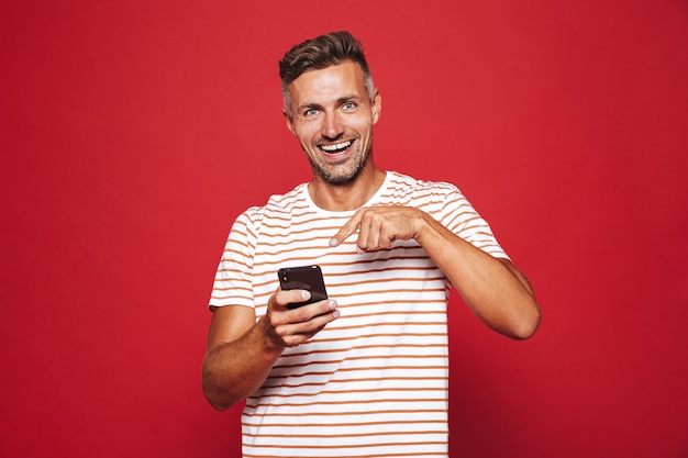 Hombre alegre de 30 años en camiseta a rayas sonriendo y sosteniendo el teléfono móvil aislado en rojo