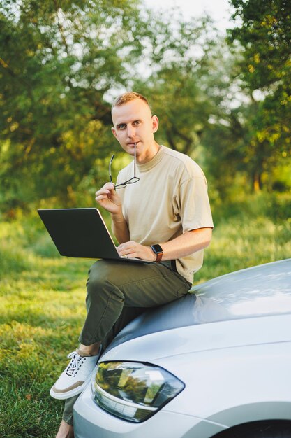 Un hombre alegre con anteojos se sienta en el capó de su automóvil en la naturaleza y trabaja en una computadora portátil Trabajo independiente trabajo remoto durante las vacaciones