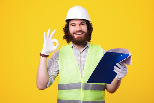 Hombre alegre arquitecto barbudo con casco y mostrando el gesto de OK mientras mira el papeleo