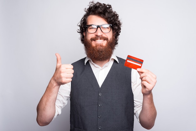 Hombre alegre barbudo sosteniendo una tarjeta de crédito y un pulgar hacia arriba sonriendo a la cámara