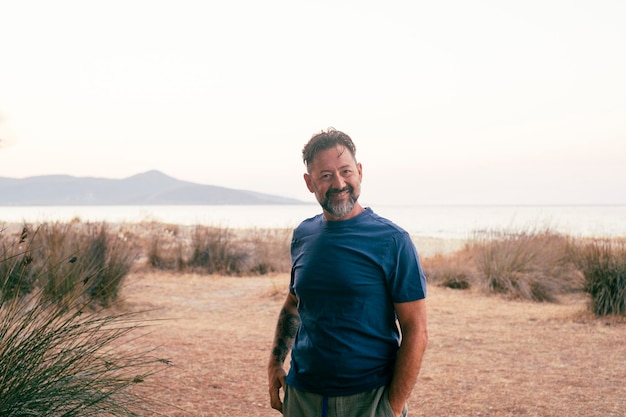 Hombre alegre caminando al aire libre feliz mirando a la cámara Fondo de destino de viaje escénico con playa e isla oceánica Espacio de copia de cielo blanco claro Ropa casual camiseta azul Estilo de vida de viaje