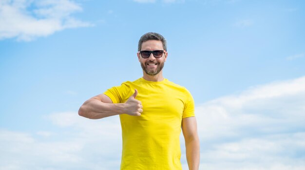 Hombre alegre con camisa amarilla y gafas de sol al aire libre en el fondo del cielo pulgar arriba