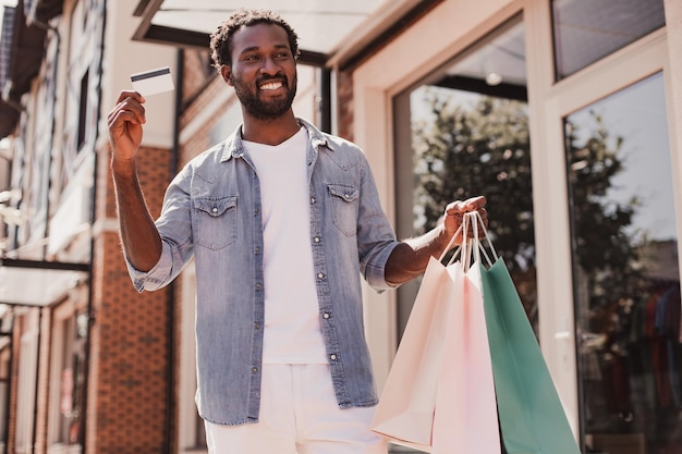 Hombre alegre con carrito de crédito en una mano y bolsas de compras en otra mirando hacia la calle