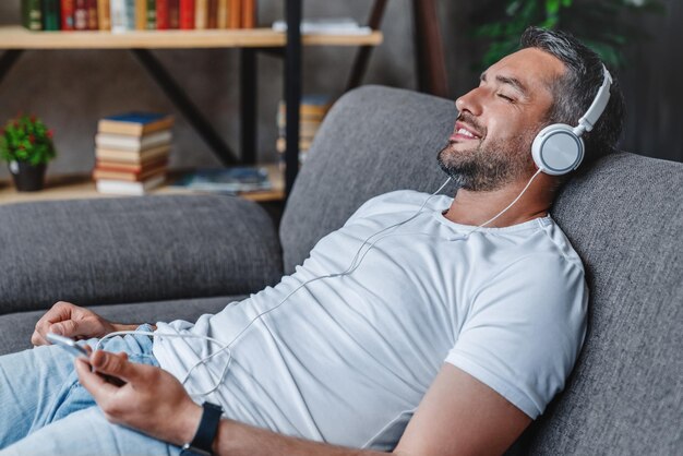 Hombre alegre en casa en el sofá escuchando música con teléfono inteligente